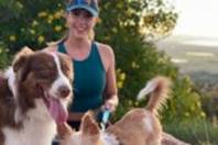 Jennifer Board looks tot he camera and smiles as she holds two dogs' leads while on a hilltop with a shrub behind her.