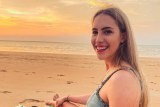 Jennifer Board smiles as she looks back to the camera while sat on the beach at what appears to be sunset.
