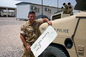 Soldier wearing camouflage gear stands next to tank holding sheet with clearly photoshopped message upon it.