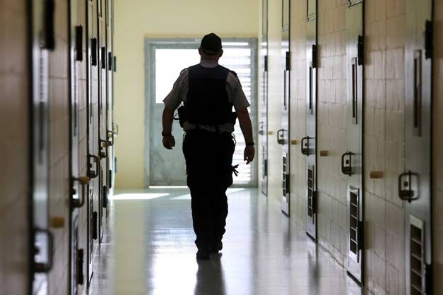 A silhouetted corrections officer walks down a cell corridor in a prison.